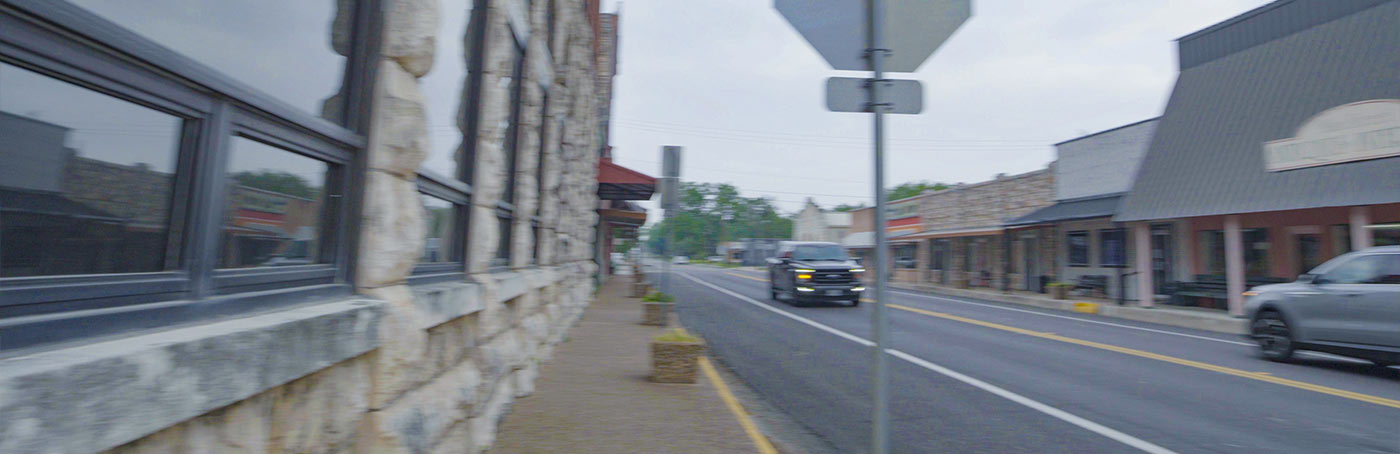 Street view showing a stone building on the left, shops on the right, a gray vehicle driving, and an overcast sky.