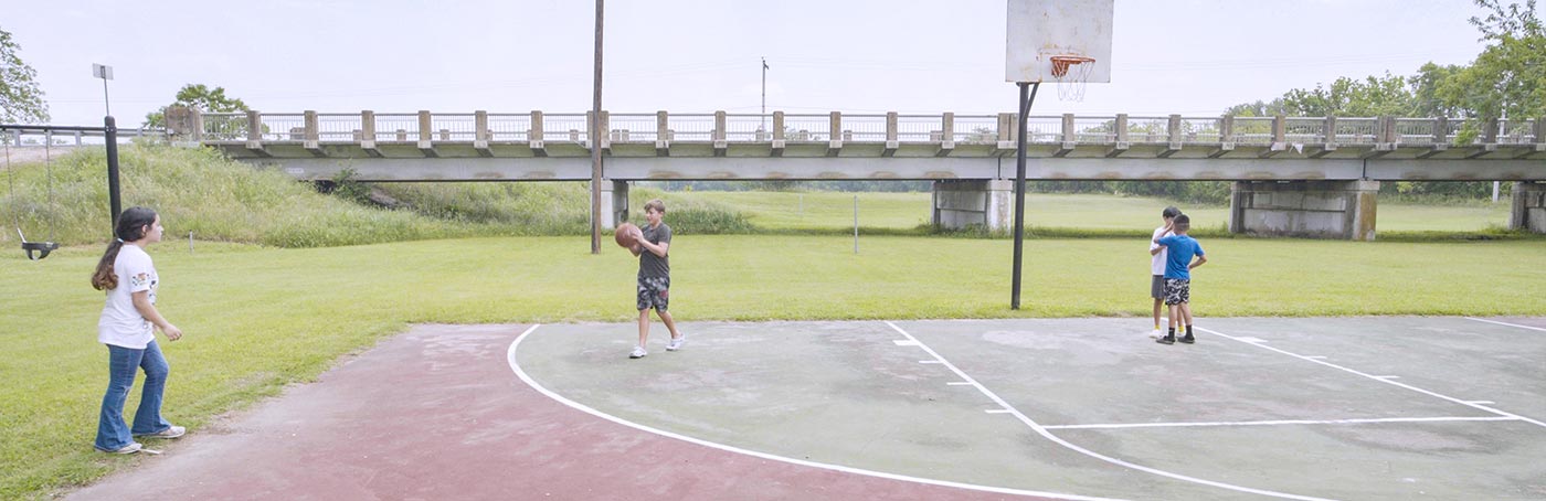 Three children play basketball on an outdoor court, with a bridge visible in the background.
