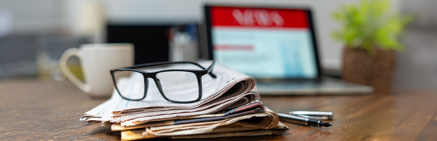 Image of glasses sitting on top of a newspaper, with an open laptop and a cup of coffee in the background